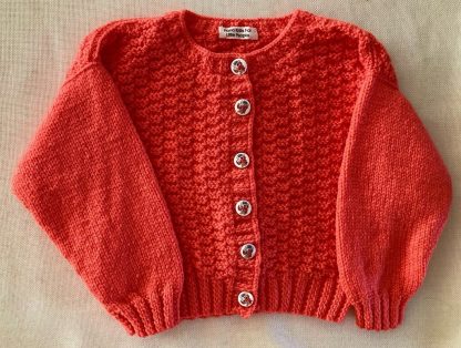 Hand knitted salmon coloured patterned cardigan Round neck 6 Delightful buttons in front Long sleeves Ribbing around neckline, cuffs and hem 100% Acrylic yarn