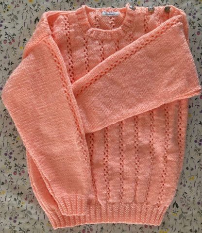 Hand knitted jumper in peach in a beautiful pattern Round neck 3 Unusual buttons on one shoulder Long sleeves Ribbing around neckline, cuffs and hem 100% Acrylic yarn Completed garment measurements Chest all around: 82cm Length, shoulder to hem ribbing: 48cm Sleeve seam length: 38cm For ages 7-8 years